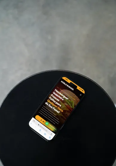 Celular apoiado numa mesa preta exibindo a home de uma hamburgueria, com o botão de pedido em destaque